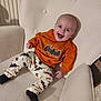 baby, child, smiling, happy, batman, orange_clothing, pants, socks, chair, armchair, cushion, indoor, person, face, cute, infant, seated, clothing, portrait, home