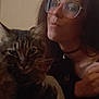 person, glasses, cat, tabby_cat, indoor, choker_necklace, brown_hair, furniture, shelf, bottle, face, pet, animal, closeup, portrait, resting, furry, whiskers, cozy, casual