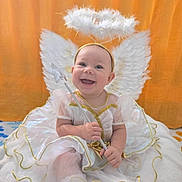 Stella is registered to the contest to win money with this photo: baby, angel, costume, wings, halo, wand, smiling, cute, white_dress, gold_trim, soft_cushion, happy, portrait, indoors, child, feathers, playful, festive, adorable, infant