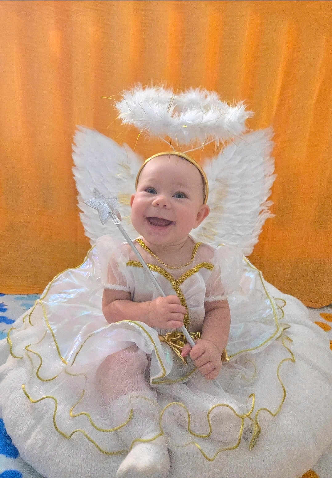 Stella is registered to the contest to win money with this photo: baby, angel, costume, wings, halo, wand, smiling, cute, white_dress, gold_trim, soft_cushion, happy, portrait, indoors, child, feathers, playful, festive, adorable, infant