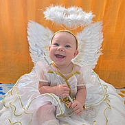 Stella is registered to the contest to win money with this photo: angel, baby, child, costume, cushion, cute, fabric, gold_trim, halo, happy, indoor, infant, orange_background, portrait, sitting, smiling, soft, wand, white_dress, wings