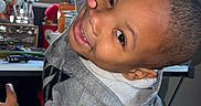 Zyon joined the competition — help win amazing prizes! child, boy, smiling, portrait, indoors, bedroom, casual_clothing, hoodie, gray_clothing, short_hair, hand_gesture, playful, happy, sitting, close_up, window, daylight, furniture, household_items, colorful_blanket