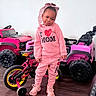 Zyair is registered to the contest to win money with this photo: child, toddler, girl, portrait, standing, smiling, pink_clothing, hoodie, sweatpants, boots, headband, toy_car, toy_truck, tricycle, bicycle, indoors, wooden_floor, playroom, vehicle, fashion