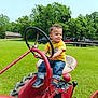 Cameron is registered to the contest to win money with this photo: toddler, child, tractor, steering_wheel, grass, field, trees, outdoor, yellow_shirt, blue_jeans, sneakers, farm_equipment, vehicle, sunny_day, nature, parked_trailers, curly_hair, small_smile, playful, rural