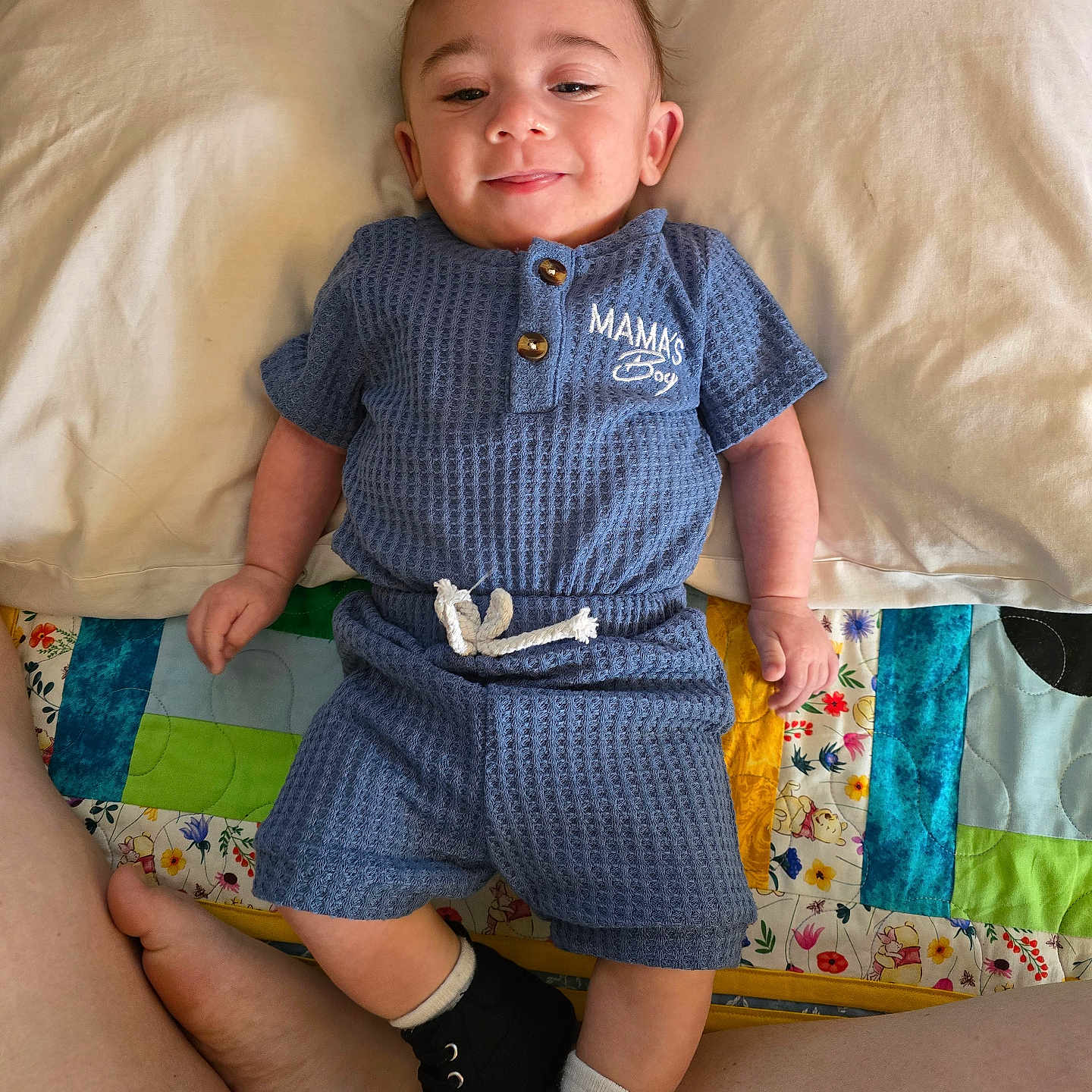 Micah is registered to the contest to win money with this photo: baby, bed, black_shoes, blue_outfit, child, cute, fabric, feet, happy, indoor, infant, legs, person, pillow, portrait, quilt, relaxed, smiling, text_on_clothing, white_socks
