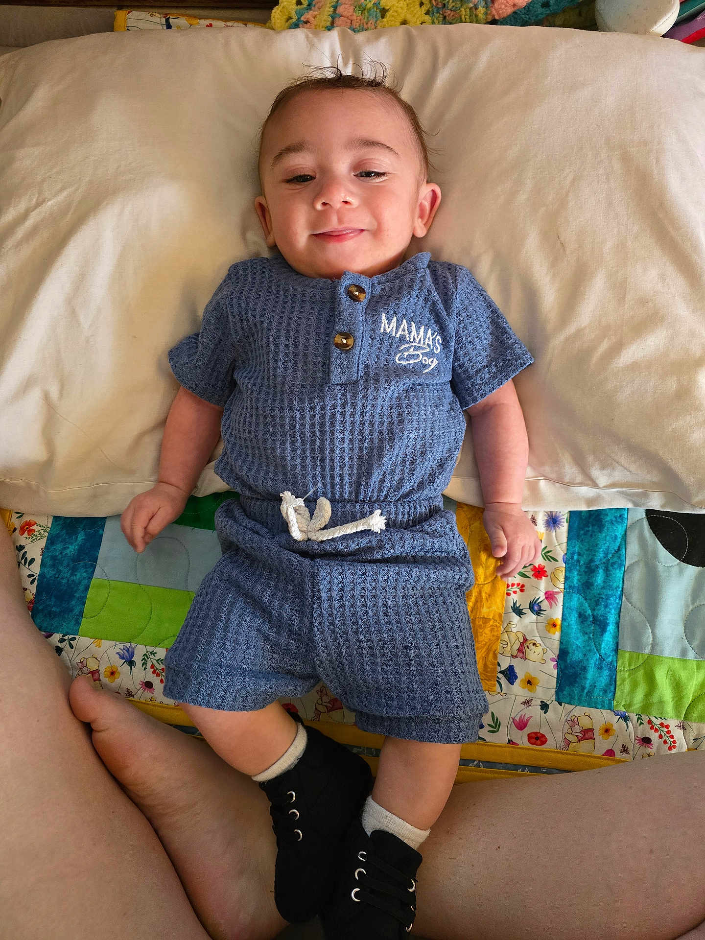 Micah is registered to the contest to win money with this photo: baby, child, smiling, blue_outfit, pillow, quilt, bed, feet, legs, black_shoes, white_socks, infant, happy, portrait, indoor, cute, person, fabric, text_on_clothing, relaxed