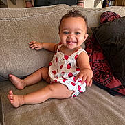 Sienna is registered to the contest to win money with this photo: baby, child, smiling, sitting, couch, romper, strawberry_pattern, happy, indoor, person, furniture, cushion, skin, foot, hand, head, face, blue_eyes, curly_hair, tattoo