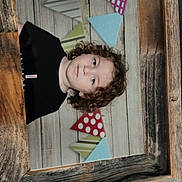 Lorenzo participe au concours pour gagner de l'argent avec cette photo : child, kid, portrait, curly_hair, wooden_frame, bunting, party_decor, background, photograph, face, eyes, neutral_expression, black_shirt, indoor, studio_style, headshot, framed_photo, wood_texture, polka_dot_pattern, cute