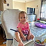 toddler, child, high_chair, pink_shirt, smiling, foot, cereal_bowl, cereal, living_room, television, curtains, table, wood_floor, toys, plastic_bottle, face, hair, seat_belt, furniture, daylight