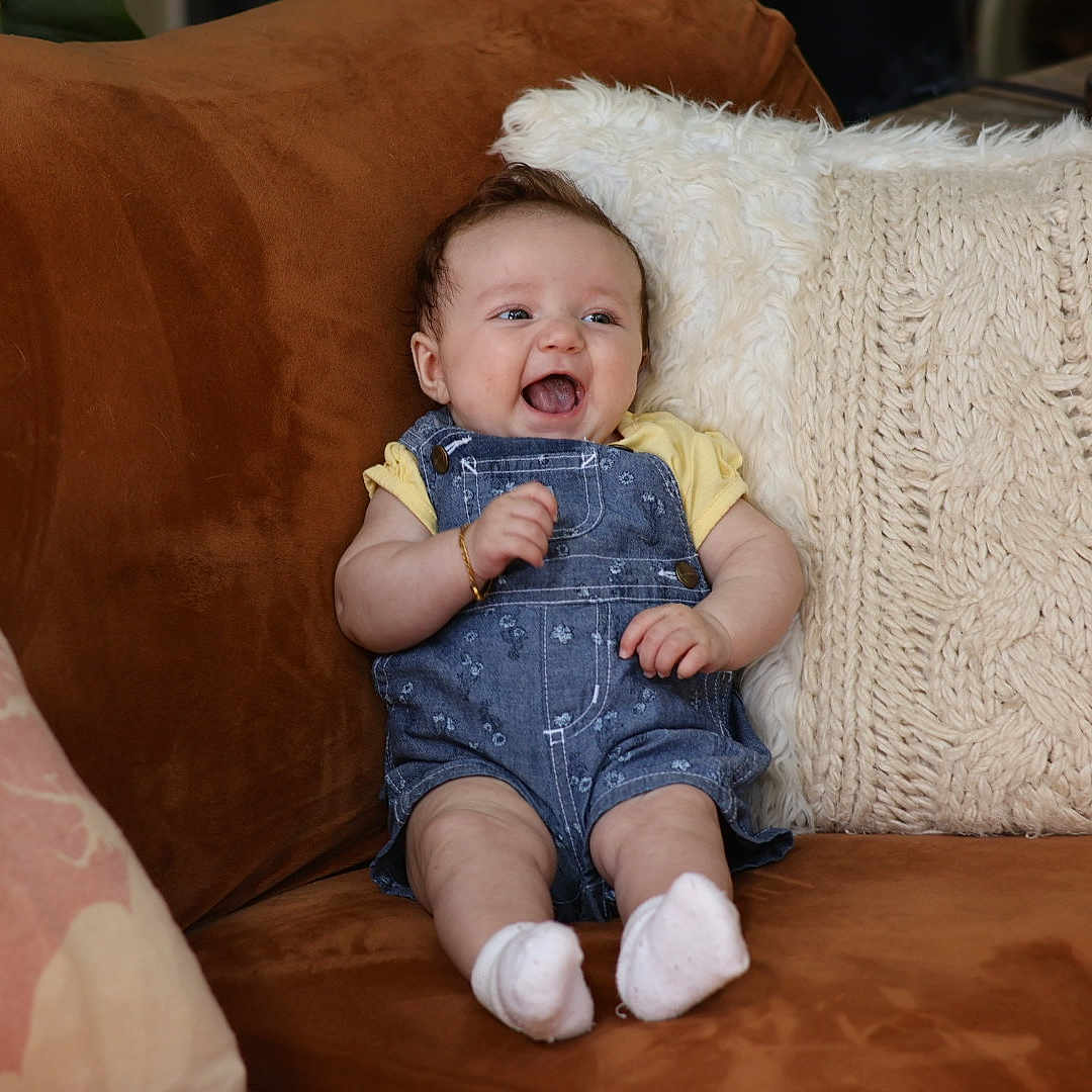 Rhaelynn is registered to the contest to win money with this photo: baby, brown_couch, child, couch, cozy, cute, denim_overalls, fluffy_pillow, happy, home, indoor, infant, knitted_pillow, person, pillow, portrait, seated, smiling, white_socks, yellow_shirt