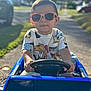 toddler, child, sunglasses, toy_car, outdoor, sunlight, street, mickey_mouse, shirt, driving, hands, steering_wheel, blue, casual_clothing, sidewalk, residential_area, daytime, cute, portrait, person