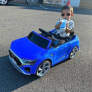 Antonio is registered to the contest to win money with this photo: toddler, child, toy_car, blue_car, sunglasses, drink, cup, outdoor, asphalt, sunlight, summer, casual_clothing, small_vehicle, parking_lot, shadow, playtime, cool_kid, car_toy, seatbelt, road