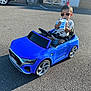 toddler, child, toy_car, blue_car, sunglasses, drink, cup, outdoor, asphalt, sunlight, summer, casual_clothing, small_vehicle, parking_lot, shadow, playtime, cool_kid, car_toy, seatbelt, road