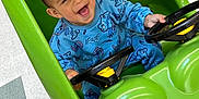 Antonio is registered to the contest to win money with this photo: toddler, child, toy_car, blue_clothing, indoor, green, happy, smiling, steering_wheel, floor_tiles, playful, cute, person, sitting, small_vehicle, joyful, casual_wear, head, face, hands