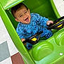 toddler, child, toy_car, blue_clothing, indoor, green, happy, smiling, steering_wheel, floor_tiles, playful, cute, person, sitting, small_vehicle, joyful, casual_wear, head, face, hands