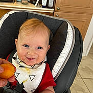 Justin is registered to the contest to win money with this photo: baby, smile, high_chair, bib, food, hand, kitchen, cabinet, orange_food, red_shirt, blue_eyes, messy, happy, infant, person, indoor, seat, cute, child, feeding