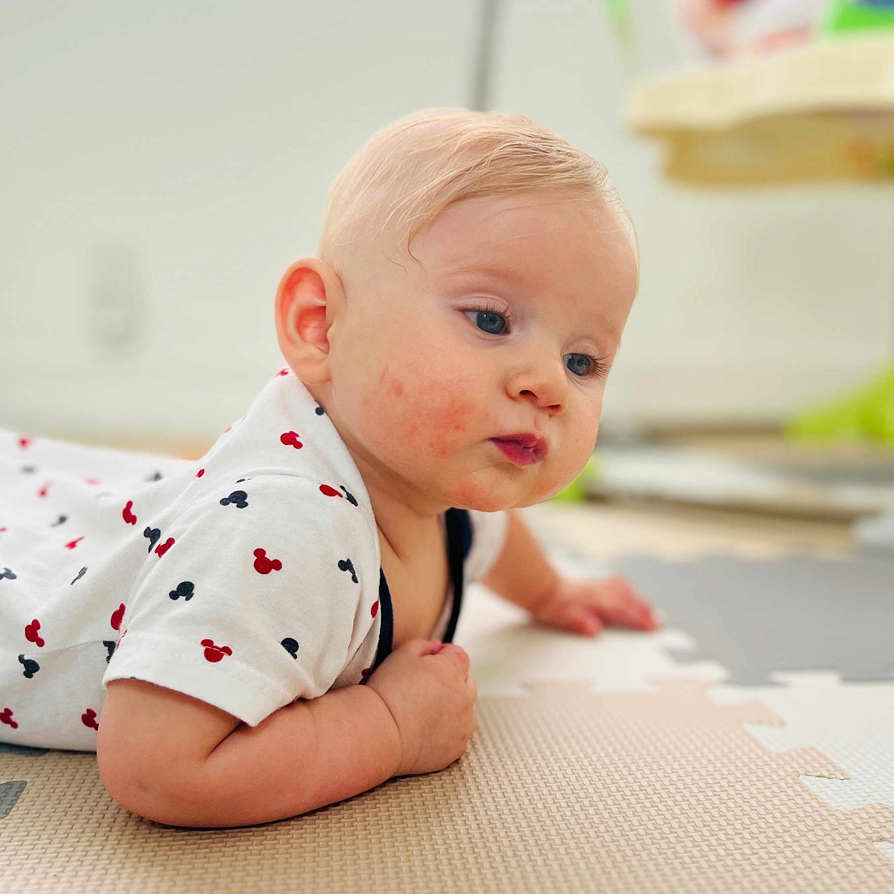 Justin is registered to the contest to win money with this photo: arm, baby, blonde_hair, blue_eyes, casual_clothing, child, curious, cute, face, floor, hand, indoor, infant, laying_down, mickey_mouse, play_mat, portrait, skin, tshirt, young_child