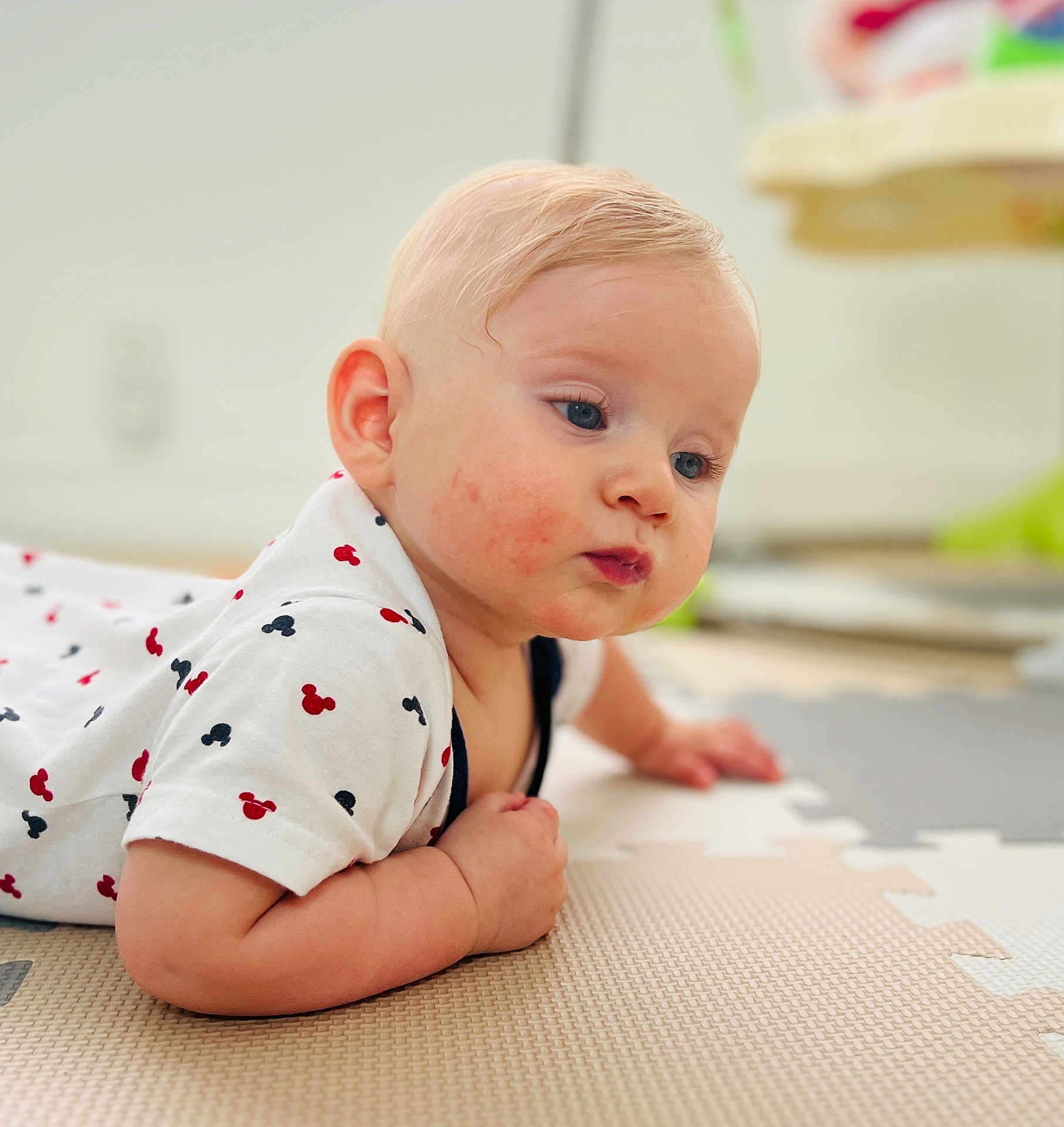 Justin is registered to the contest to win money with this photo: baby, infant, child, blonde_hair, blue_eyes, tshirt, mickey_mouse, play_mat, floor, curious, cute, indoor, skin, face, hand, arm, young_child, casual_clothing, portrait, laying_down