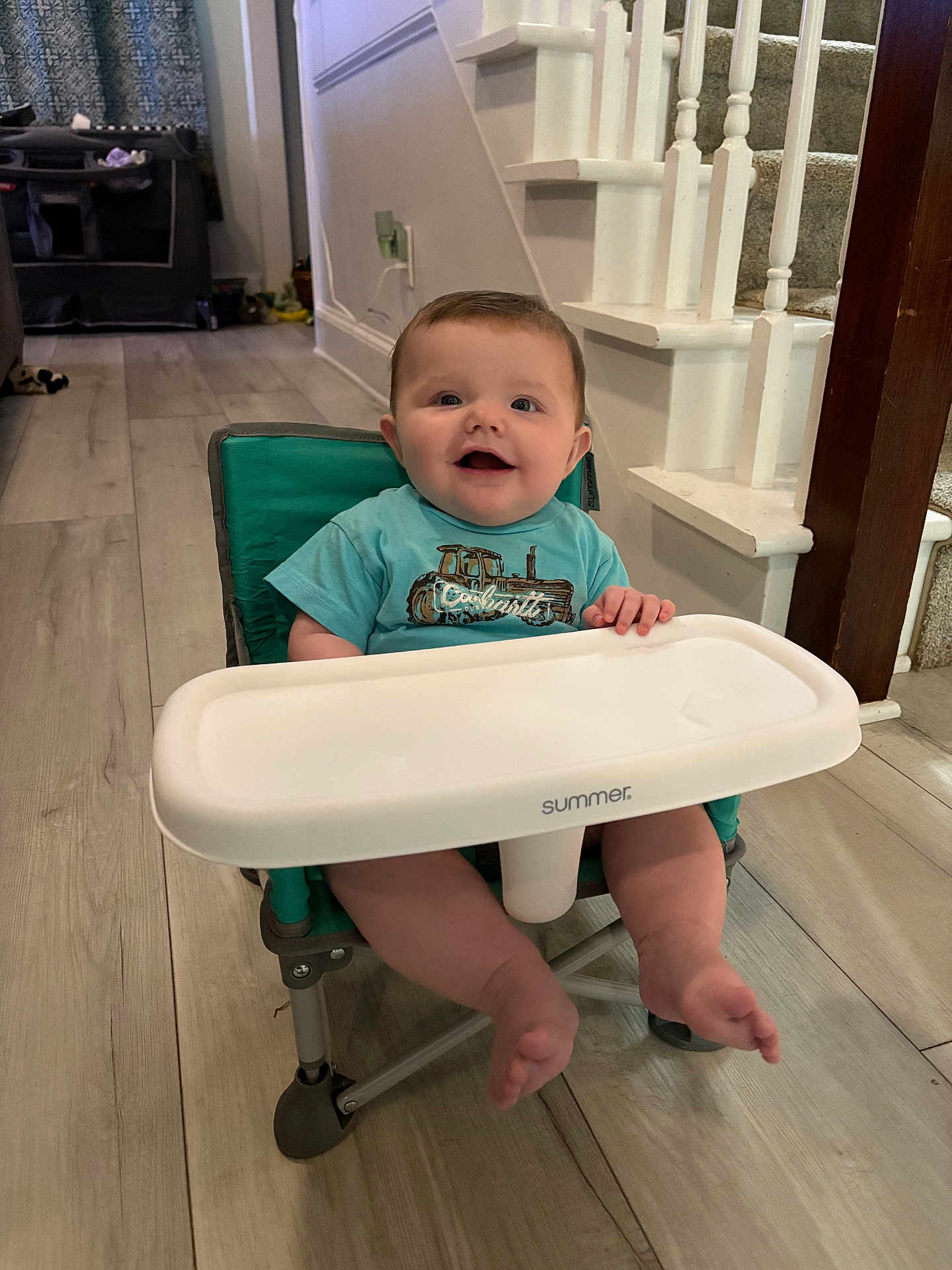 Lilith is registered to the contest to win money with this photo: baby, child, high_chair, smiling, blue_shirt, indoor, wooden_floor, staircase, home, person, face, tray, foot, hand, furniture, happy, flooring, infant, sitting, casual