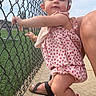 toddler, child, pink_sandals, floral_outfit, chain_link_fence, sidewalk, outdoor, daylight, person, footwear, grass, sky, hair_bow, bracelet, smile, holding, confident, cute, summer, casual
