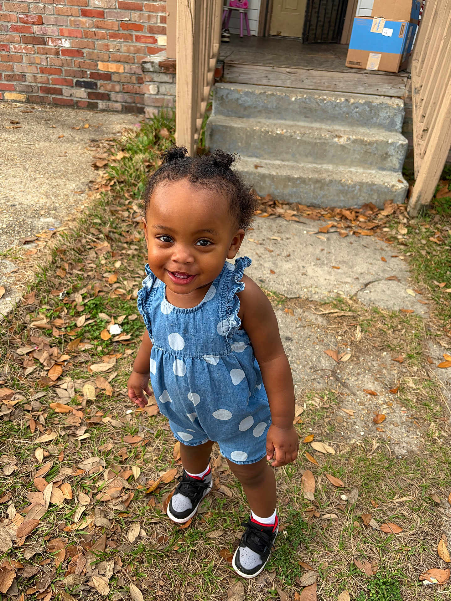 Reign is registered to the contest to win money with this photo: toddler, child, outdoor, grass, leaves, steps, porch, brick_wall, concrete, romper, polka_dot, smiling, hair_buns, sneakers, white_socks, happy, person, young_child, casual_clothing, daylight