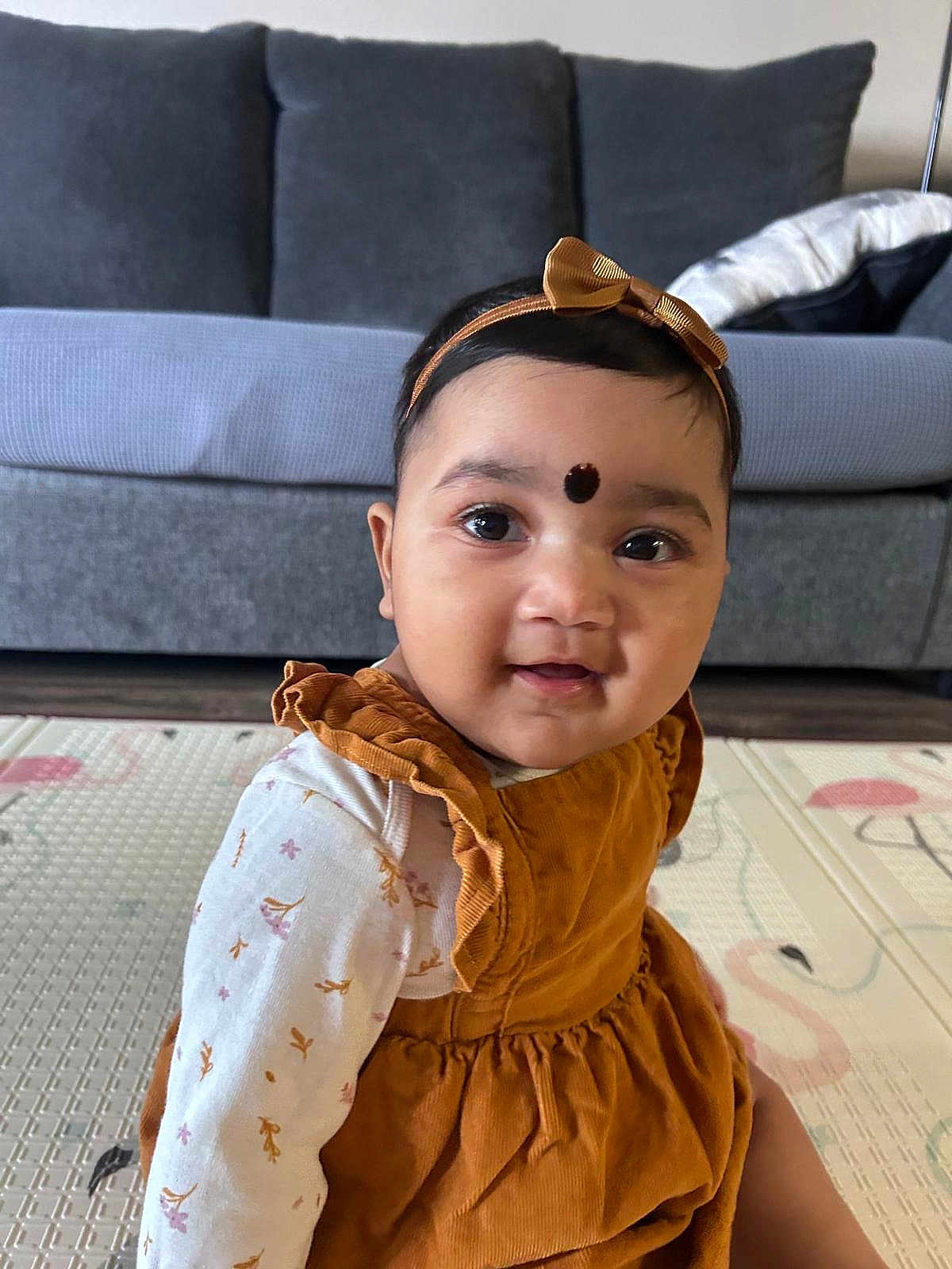Manasvi is registered to the contest to win money with this photo: baby, bow, child, clothing, couch, cute, dress, face, floor, girl, happy, headband, home, indoors, long_sleeve, mat, pattern, portrait, sitting, smile