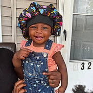 Skyler is registered to the contest to win money with this photo: toddler, child, smiling, denim_overalls, bonnet, baby, person, hand, door, house_number, outdoor, portrait, cute, happy, skin, clothing, holding, face, window, family