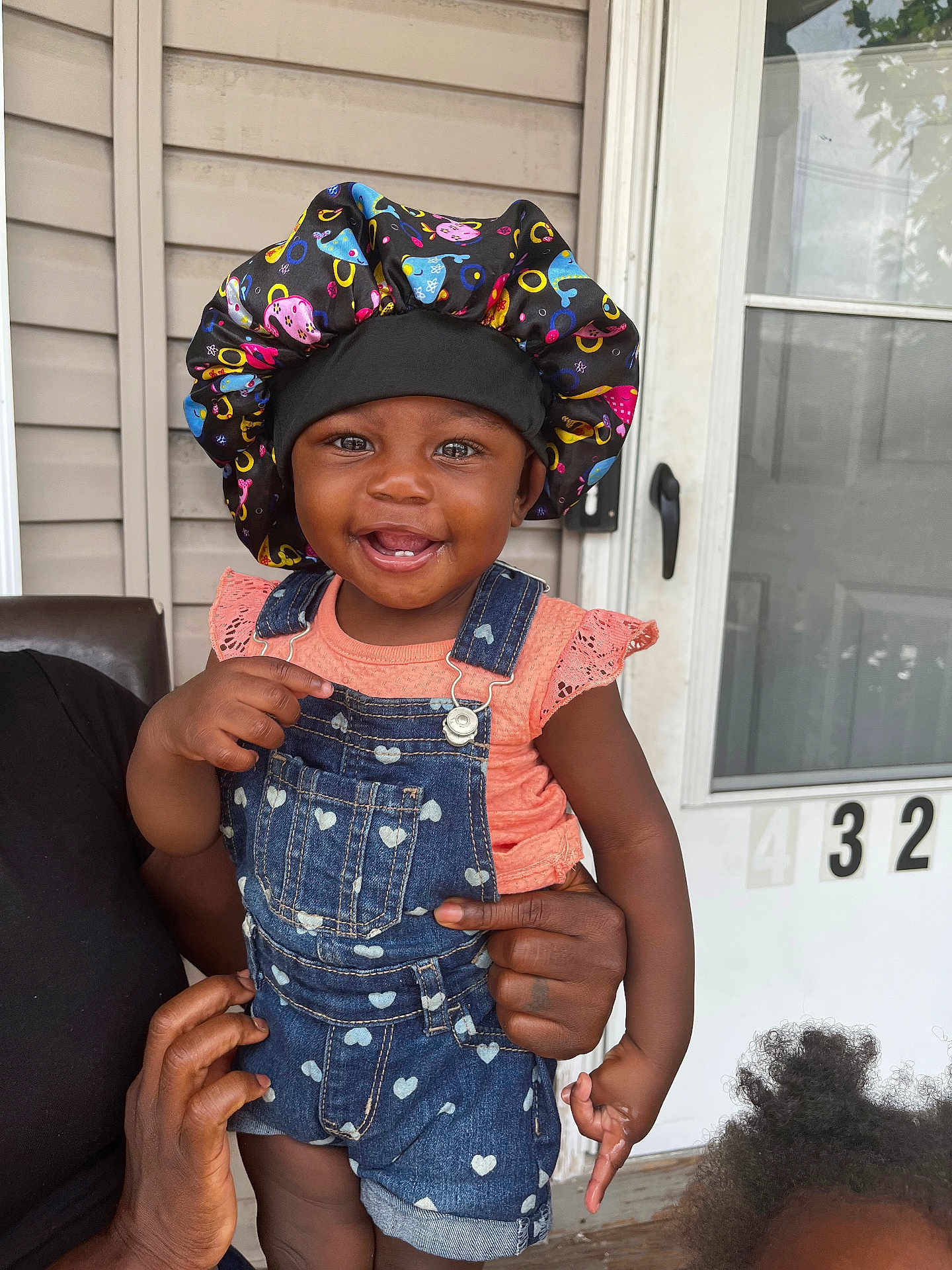 Skyler is registered to the contest to win money with this photo: toddler, child, smiling, denim_overalls, bonnet, baby, person, hand, door, house_number, outdoor, portrait, cute, happy, skin, clothing, holding, face, window, family