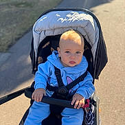 Leo is registered to the contest to win money with this photo: baby, child, stroller, blue_clothing, hoodie, outdoor, sunlight, sitting, handrail, pathway, grass, infant, cute, daylight, baby_face, baby_hands, safety_harness, casual_wear, young_child, portrait