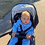 baby, child, stroller, blue_clothing, hoodie, outdoor, sunlight, sitting, handrail, pathway, grass, infant, cute, daylight, baby_face, baby_hands, safety_harness, casual_wear, young_child, portrait