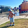 Anthony joined the competition — help win amazing prizes! toddler, child, blue_cap, shorts, shoes, grass, water_play_area, sunny_day, outdoor, playground, children, water, fence, sidewalk, people, summer, fun, park, hat, curious