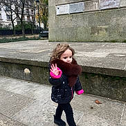 Esther a rejoint le concours — aidez-le/la à gagner de superbes lots ! child, walking, winter_clothing, jacket, fur_collar, black_shoes, sidewalk, stone_wall, urban, trees, autumn, waving_hand, curly_hair, outdoor, pavement, casual, person, cold_weather, young_child, fashion