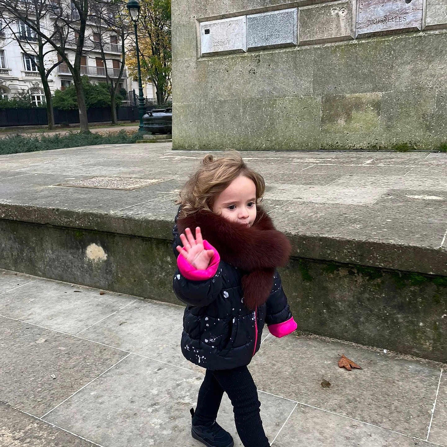 Esther a rejoint le concours — aidez-le/la à gagner de superbes lots ! autumn, black_shoes, casual, child, cold_weather, curly_hair, fashion, fur_collar, jacket, outdoor, pavement, person, sidewalk, stone_wall, trees, urban, walking, waving_hand, winter_clothing, young_child