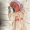 baby, bed, blanket, bottle_nipple, cozy, eyes_open, face, floral_clothing, indoors, infant, lying_down, mittens, newborn, onesie, pacifier, portrait, sleepwear, striped_cap, textile, white_bedding
