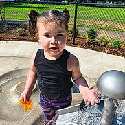 Melinda is registered to the contest to win money with this photo: child, toddler, water_play, outdoor, playground, sunlight, water_toy, pigtails, black_tank_top, colorful_pants, pink_sandals, water_splash, concrete_surface, metal_fountain, green_grass, chain_link_fence, trees, daytime, curious_expression, park
