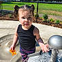 child, toddler, water_play, outdoor, playground, sunlight, water_toy, pigtails, black_tank_top, colorful_pants, pink_sandals, water_splash, concrete_surface, metal_fountain, green_grass, chain_link_fence, trees, daytime, curious_expression, park