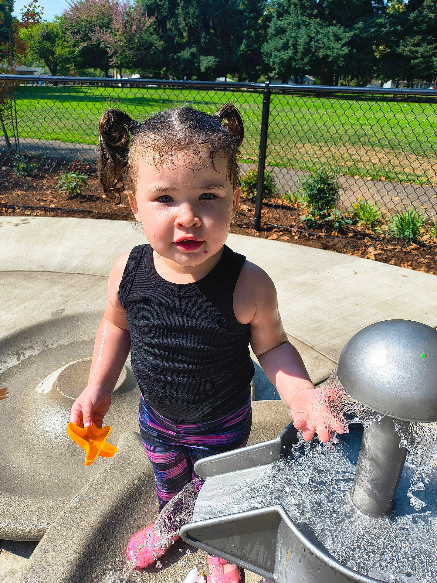 Melinda is registered to the contest to win money with this photo: child, toddler, water_play, outdoor, playground, sunlight, water_toy, pigtails, black_tank_top, colorful_pants, pink_sandals, water_splash, concrete_surface, metal_fountain, green_grass, chain_link_fence, trees, daytime, curious_expression, park