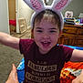 Melinda is registered to the contest to win money with this photo: arms_outstretched, blanket, bunny_ears, carpet, child, cozy, cute, family_photos, happy, home, indoor, living_room, maroon_shirt, person, playful, short_hair, sitting, smiling, toddler, wooden_furniture