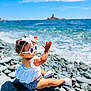 toddler, child, beach, rocks, ocean, waves, sunlight, sunglasses, white_top, checkered_shorts, headband, bow, summer, playful, outdoor, water, sky, vacation, cute, sunny