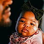 baby, child, infant, headband, bow, floral_bib, pink_clothing, face, portrait, adult_hand, blurred_adult, close_up, warm_lighting, indoor, soft_focus, skin, cute, expression, family, love