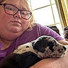 Smoky Grey is registered to the contest to win money with this photo: person, glasses, puppy, dog, indoor, window, curtains, purple_shirt, couch, arm, resting, closeup, face, sleeping_dog, casual, home, relaxation, light, comfort, pet