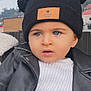 toddler, child, blue_eyes, black_hat, pom_poms, super_star_patch, white_sweater, black_jacket, outdoor, portrait, curious_expression, winter_clothing, face, headwear, cute, young_child, fashion, person, close_up, casual