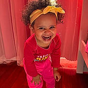 Amely is registered to the contest to win money with this photo: toddler, child, smiling, headband, curly_hair, pink_clothing, sneakers, indoor, wooden_floor, curtains, happy, person, cute, playful, young_child, standing, bright_colors, portrait, fun, warm_lighting