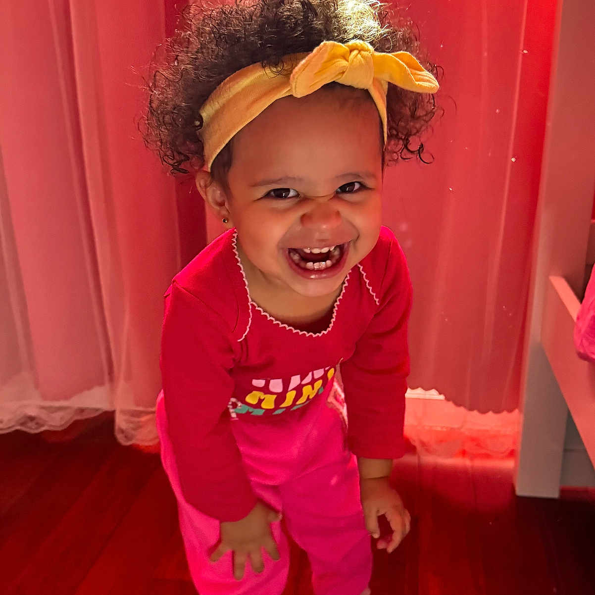 Amely is registered to the contest to win money with this photo: bright_colors, child, curly_hair, curtains, cute, fun, happy, headband, indoor, person, pink_clothing, playful, portrait, smiling, sneakers, standing, toddler, warm_lighting, wooden_floor, young_child