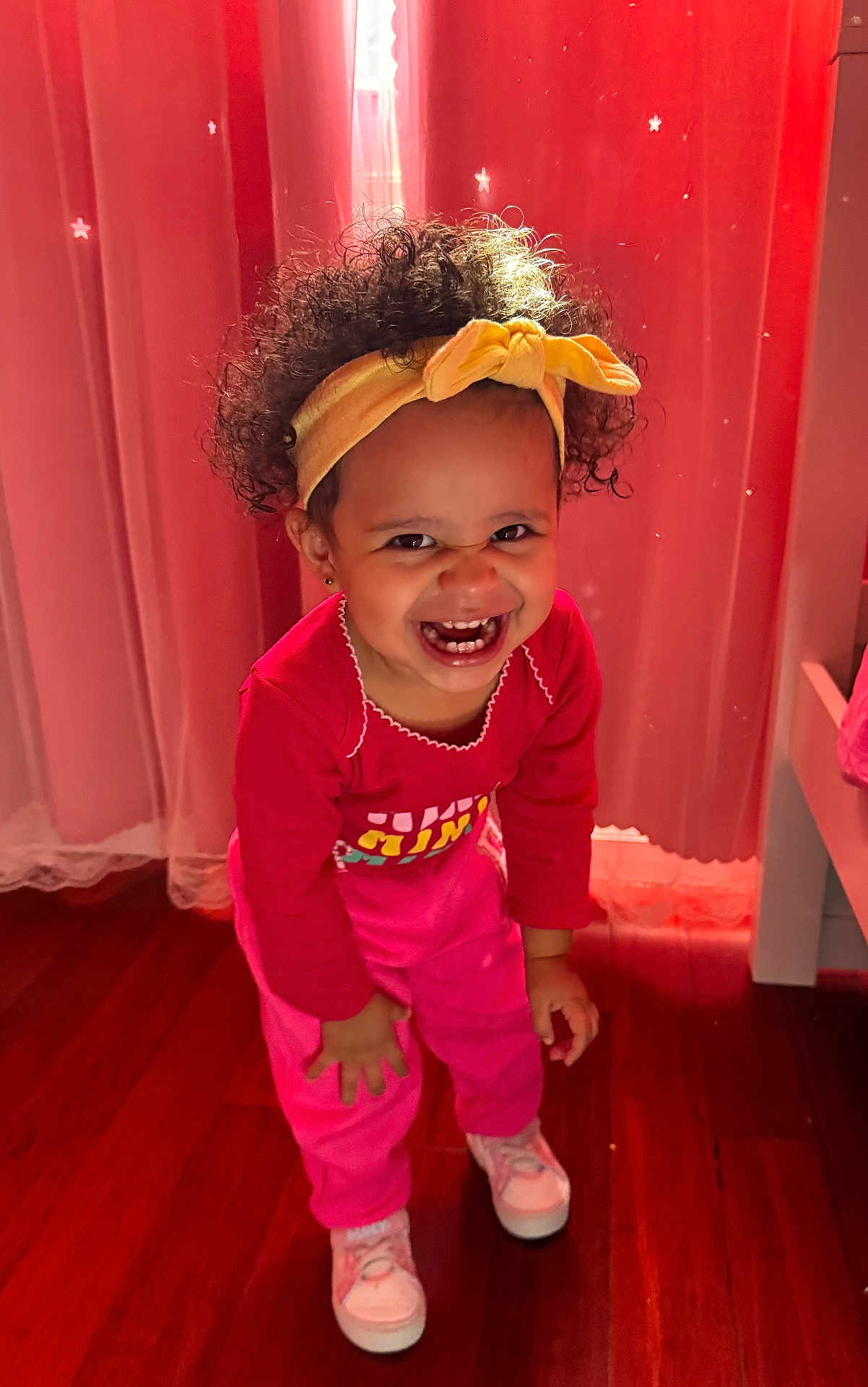 Amely is registered to the contest to win money with this photo: toddler, child, smiling, headband, curly_hair, pink_clothing, sneakers, indoor, wooden_floor, curtains, happy, person, cute, playful, young_child, standing, bright_colors, portrait, fun, warm_lighting