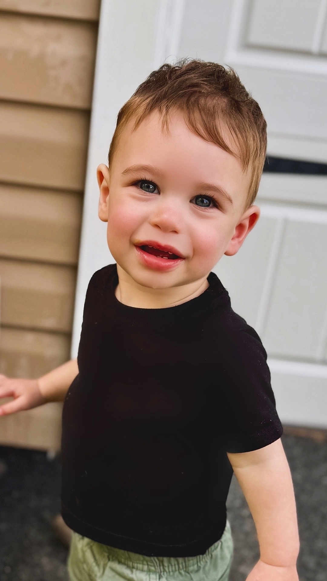 Jack is registered to the contest to win money with this photo: toddler, child, smile, black_shirt, green_shorts, short_hair, outdoor, door, house, skin, face, eyes, person, standing, happy, young_child, casual_clothing, cheerful, daylight, portrait