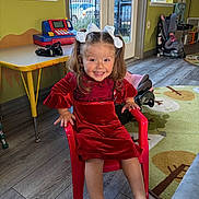 Juliana is registered to the contest to win money with this photo: child, girl, red_dress, smiling, chair, indoor, toy, playroom, hair_bow, flooring, window, table, footwear, car_seat, rug, furniture, light, happy, person, decor
