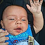 baby, infant, child, face, hand, blue_bib, black_clothing, smiling, raising_hand, close_up, skin, eyes, mouth, fingers, seat, pacifier_clip, expression, person, cute, portrait