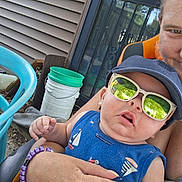 Johnny is registered to the contest to win money with this photo: baby, child, sunglasses, cap, blue_shirt, sailboat_print, adult_hand, outdoor, house_siding, bucket, chair, smile, reflection, person, holding, summer, casual, portrait, headwear, sun_protection