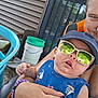 baby, child, sunglasses, cap, blue_shirt, sailboat_print, adult_hand, outdoor, house_siding, bucket, chair, smile, reflection, person, holding, summer, casual, portrait, headwear, sun_protection